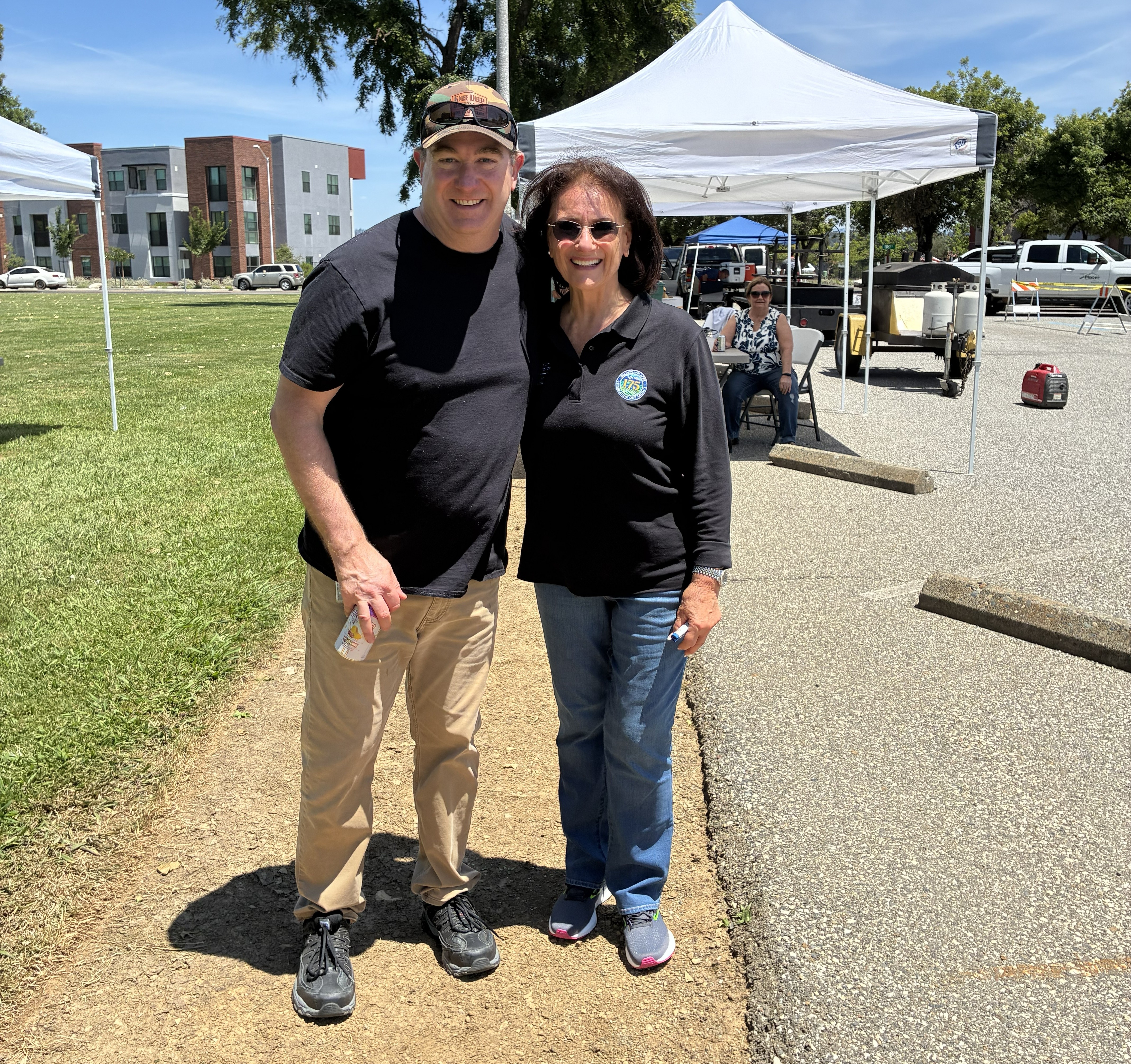 Supervisor Suzanne Jones standing with Matt Randall outside under a blue sky next to green grass in front of a white pop-up tent