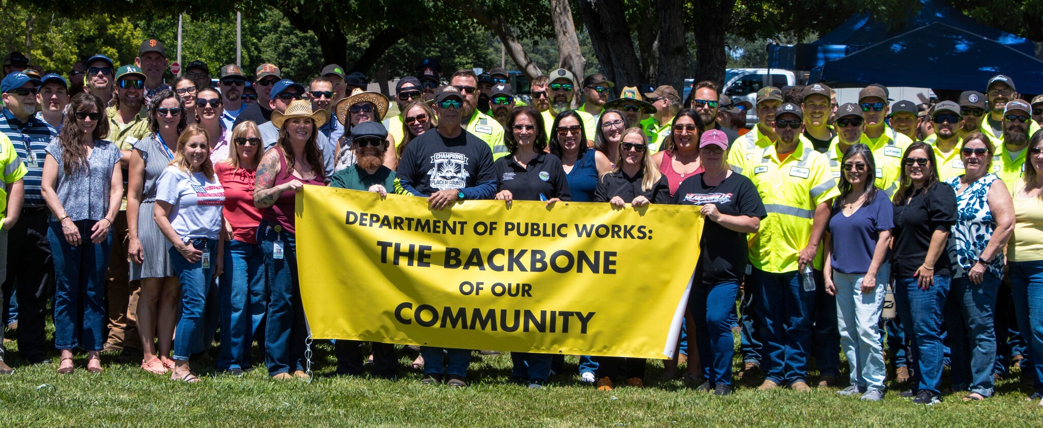 Crowd of community leaders and members of the Department of Public Works standing in green grass holding a yellow sign that reads "Department of Public Works: The Backbone of our Community"
