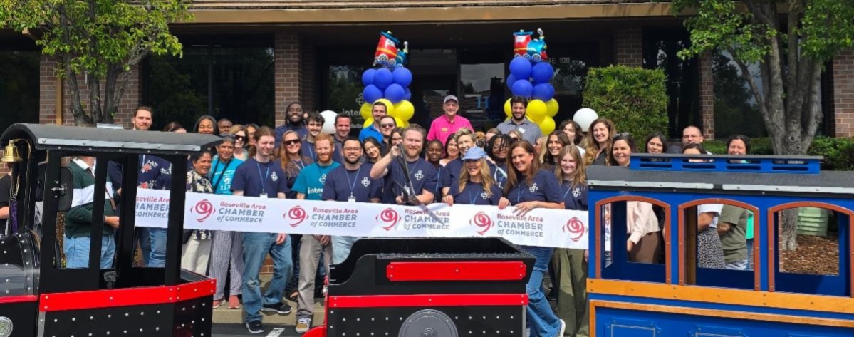 a group of people standing behind a ribbon that has the Roseville Area Chamber of Commerce logo on it behind a small colorful train at the foot of some steps to the doorway of an office building
