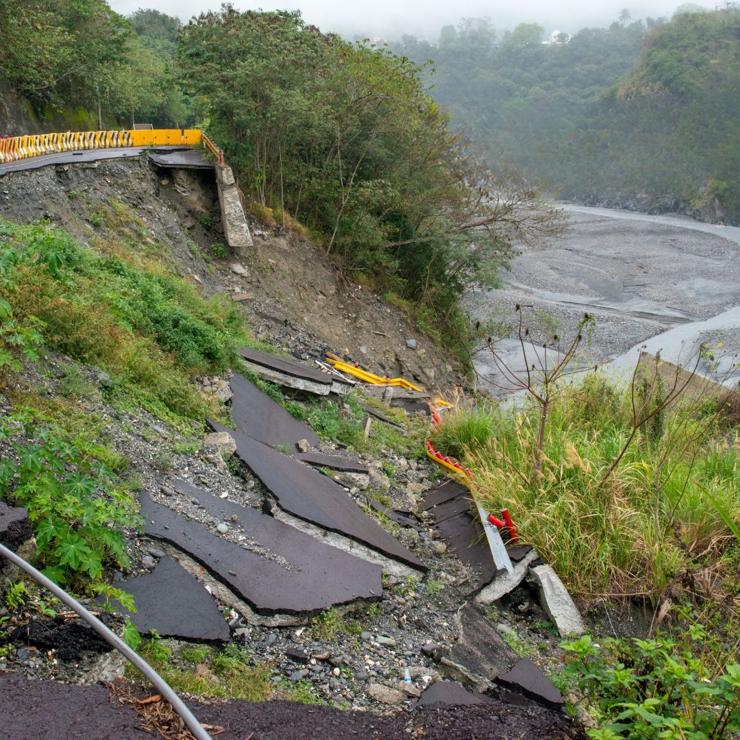A damaged hillside road with broken pavement slabs scattered down the slope. A section of the road above is partially collapsed, with yellow and red safety barriers placed near the damaged area. Dense vegetation surrounds the hillside, and a wide, rocky riverbed is visible below. Mist and distant greenery appear in the background.