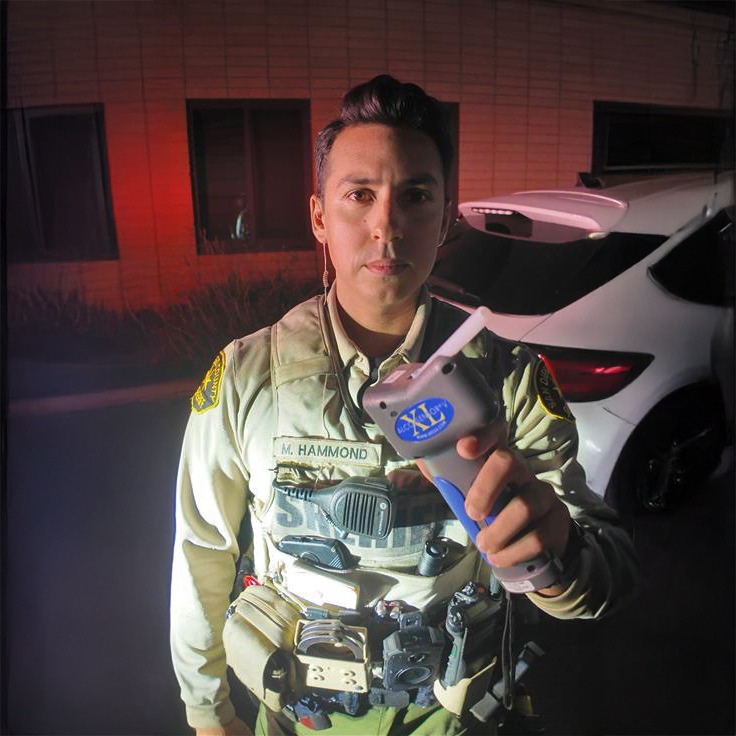 A person taking a breathalizer test to see alcohal level. shows a car in the back with a machine being held with blue sticker with yellow xl type.