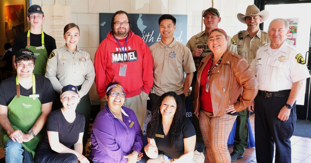 A group of people standing and kneeling indoors in a well‑lit restaurant or café setting. The group includes individuals wearing uniforms with badges, individuals wearing casual clothing, and individuals wearing aprons typically worn by food‑service workers. They are arranged in two rows, with some standing in the back and others kneeling in the front. Behind them are tiled walls, signage, and a doorway.