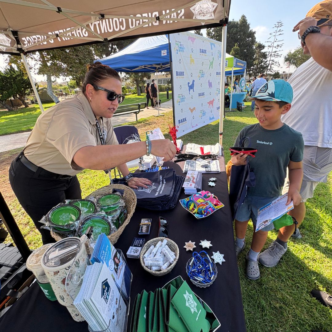 A person wearing a uniform shirt and sunglasses is standing under a canopy labeled “San Diego County Sheriff.” The person is reaching across a table filled with various giveaway items, including brochures, small containers, keychains, and star-shaped items. A child holding a phone and a flyer is standing near the table, with another adult nearby. Multiple booths and canopies are set up in the background on a grassy park area, with people walking and trees visible.