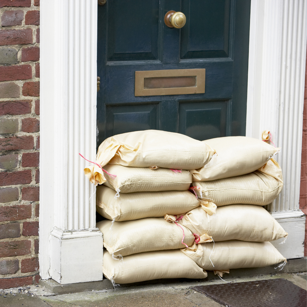 A stack of sandbags placed in front of a dark-colored door to protect a building entrance from flooding. The door is framed by white trim and set in a brick wall.