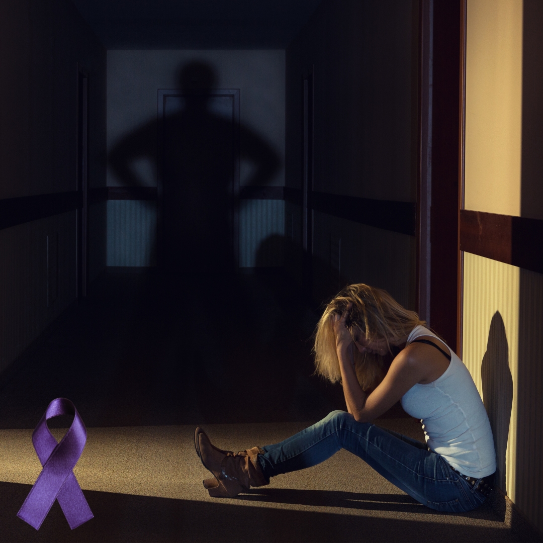 A person sitting on the floor of a dim hallway with their head down, while the shadow of another figure appears at the far end of the hallway with purple ribbon.