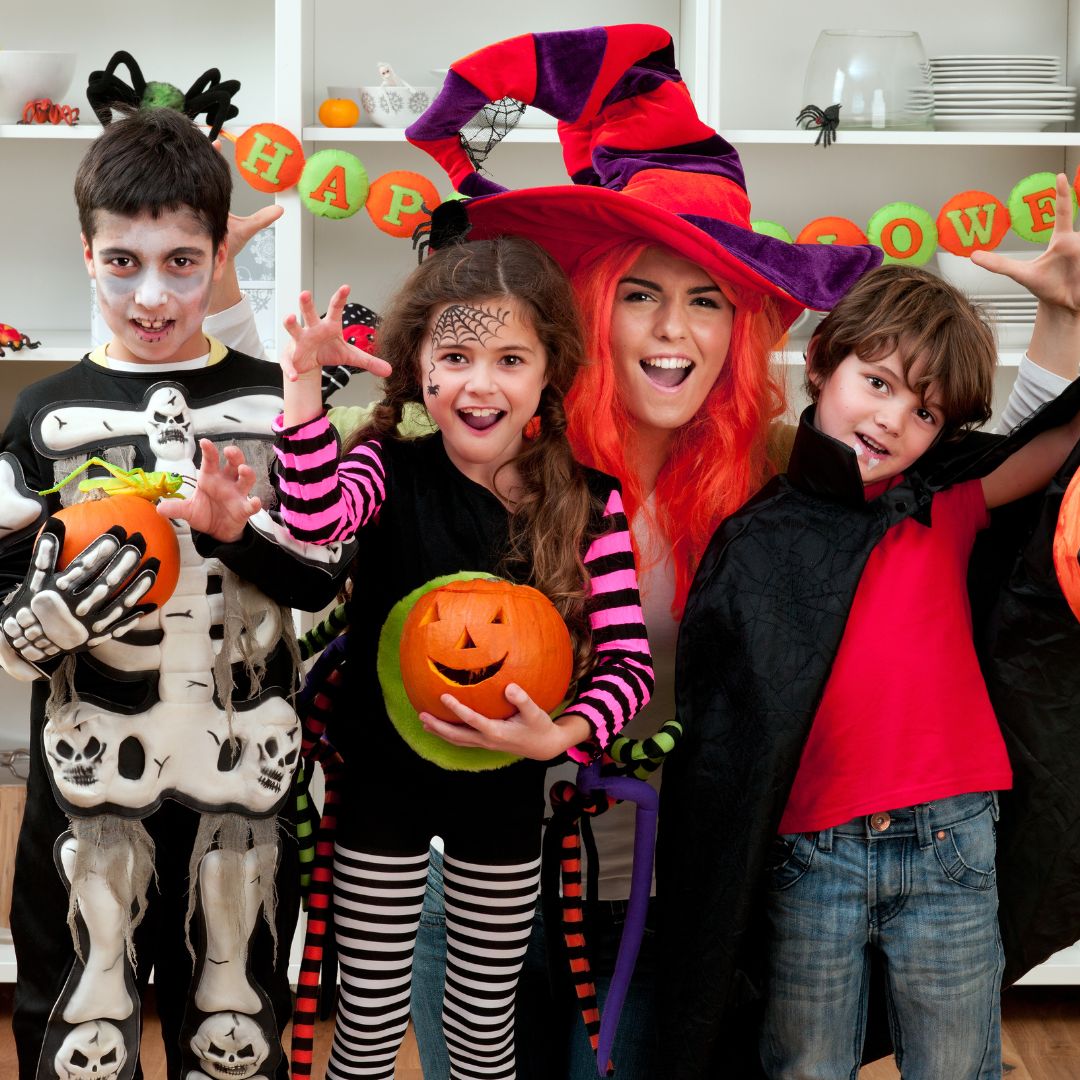Four individuals wearing Halloween costumes stand indoors in front of festive decorations. One individual is dressed in a skeleton outfit and holding a carved pumpkin. Another individual is wearing striped clothing with face paint and holding a pumpkin bucket. A third individual in the back is wearing a large hat with bright colors and a wig. The fourth individual is dressed in a cape. A “Happy Halloween” banner and shelves with decorations are visible in the background.