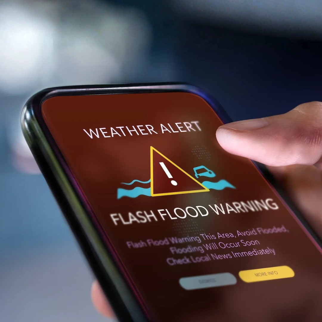 A close-up view of a smartphone displaying a weather alert screen that reads “Flash Flood Warning,” with a triangular warning icon and water graphic. A hand is touching the screen as the alert details appear below.