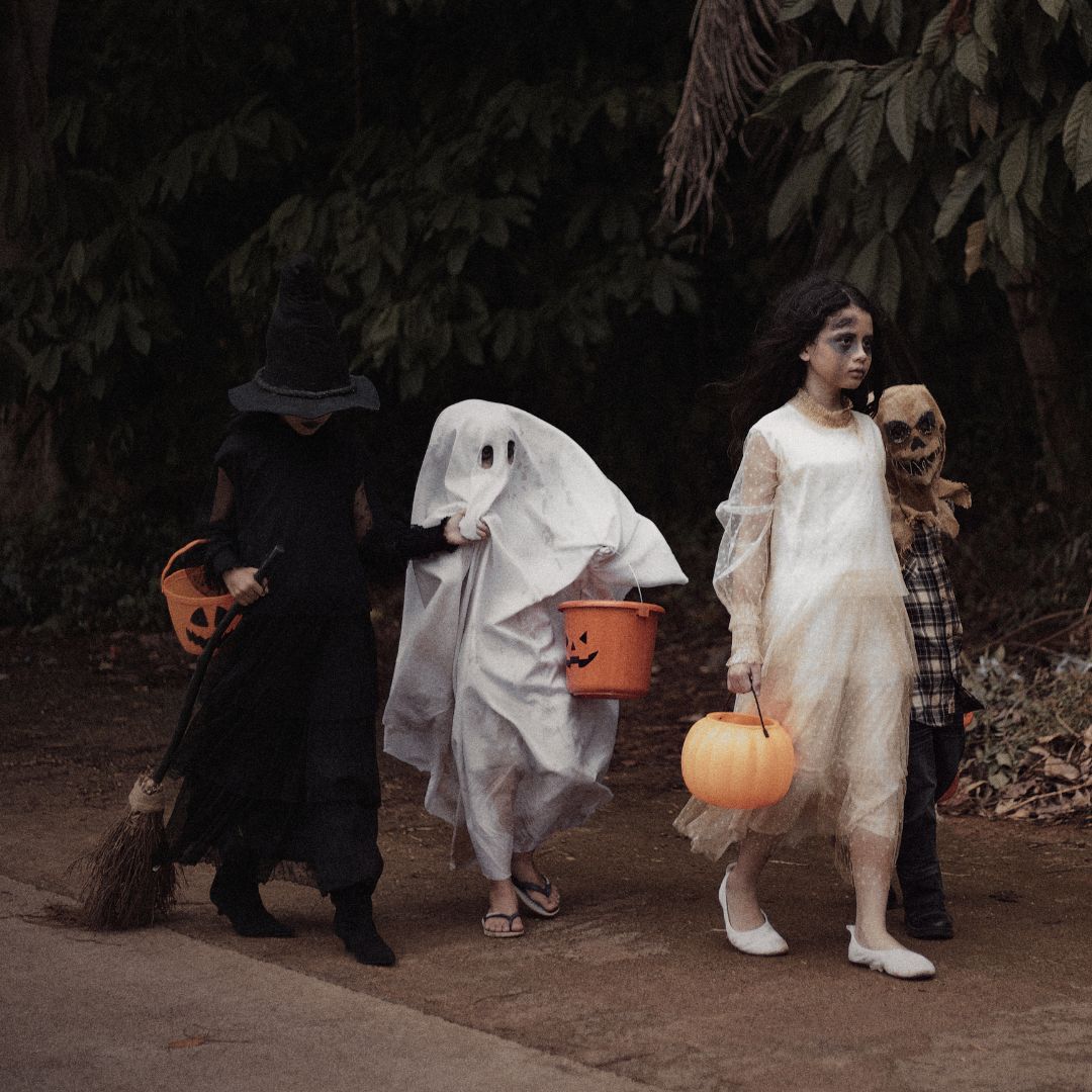 Four individuals in costumes walk outdoors along a path with trees and foliage in the background. One individual is wearing a long dark outfit and holding a broom, another is covered in a white sheet resembling a ghost, another is wearing a light-colored outfit and holding a pumpkin-shaped bucket, and the fourth is wearing a mask and patterned clothing. Each carries a pumpkin bucket for collecting treats.