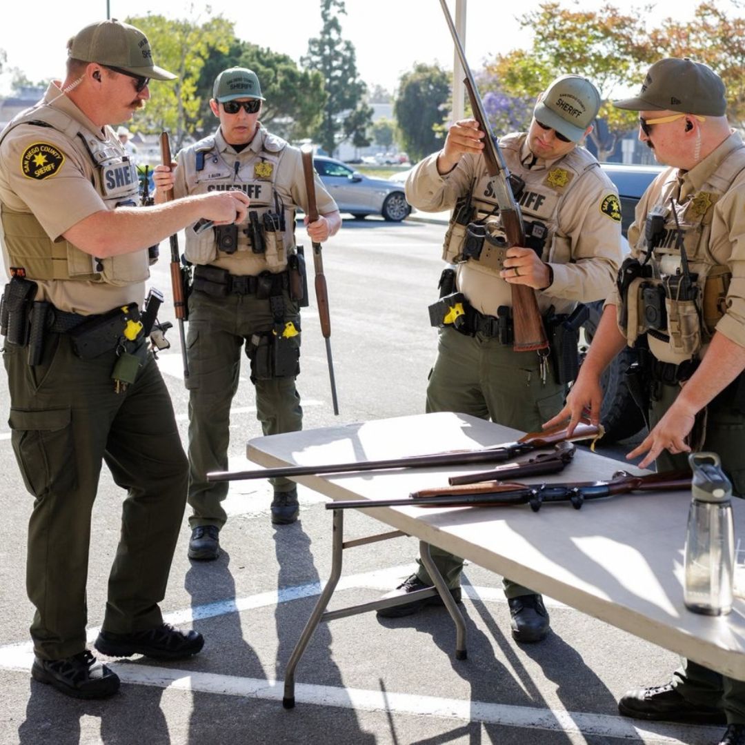A group of uniformed individuals is standing outdoors near a folding table in a paved area. Several long, dark‑colored objects are placed on the table. One individual is holding one of the objects upright, while others observe or handle similar items. Trees, parked vehicles, and buildings are visible in the background under natural daylight.