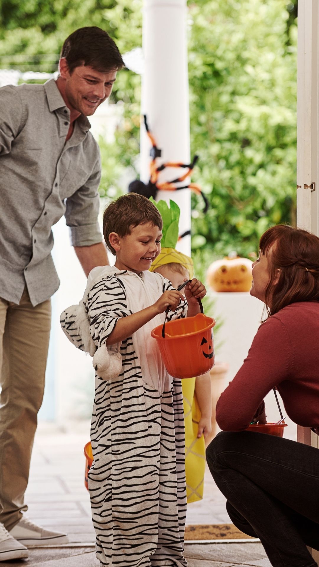 A child wearing a striped costume stands at a doorway holding an orange pumpkin-shaped bucket. Another child in a costume is partially visible behind. An adult standing nearby is leaning slightly forward toward the child, and another adult stands behind the children. The scene takes place outdoors near a doorway decorated with Halloween items, with greenery visible in the background.