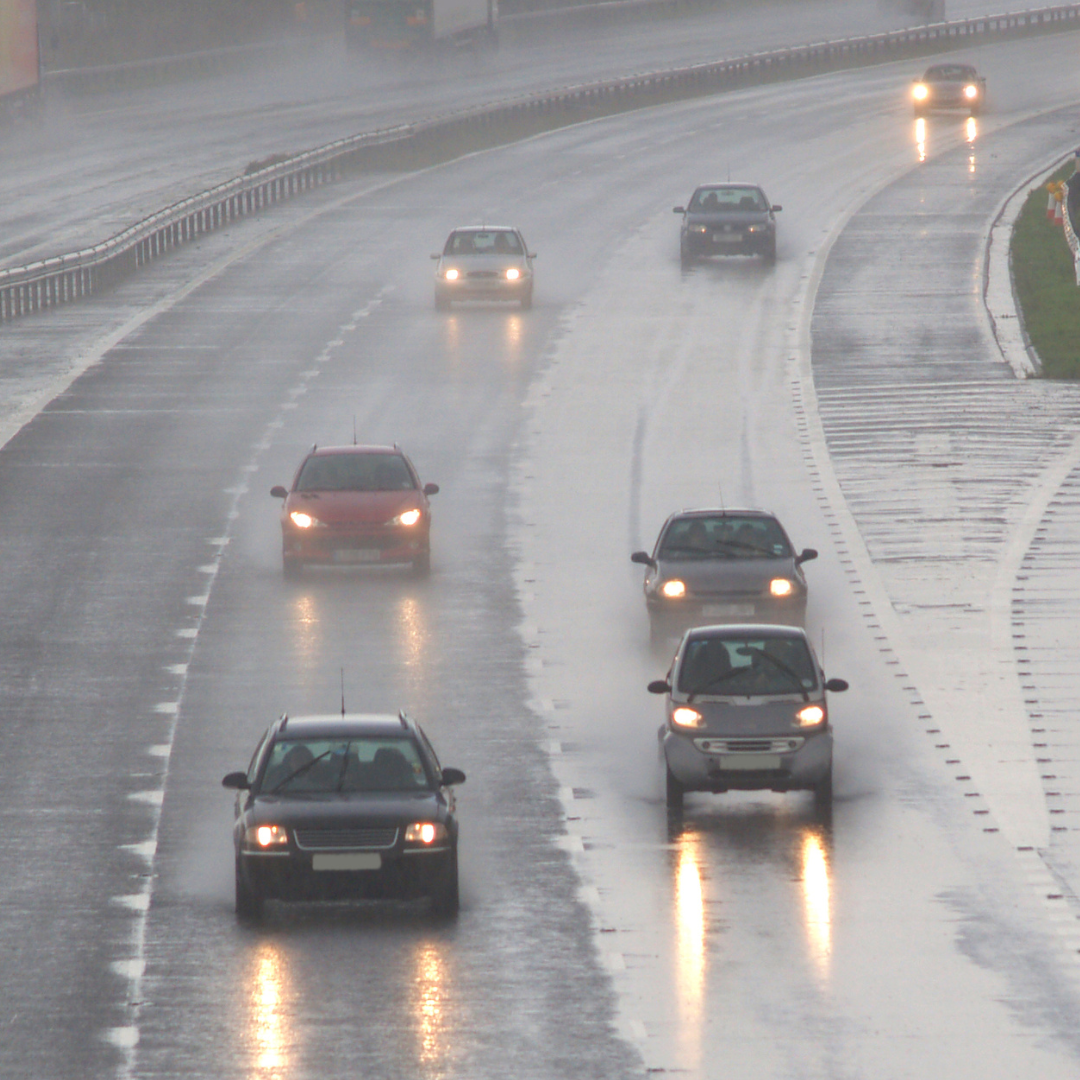 Cars driving on a wet highway during heavy rain, with headlights reflecting off the shiny road surface. Multiple lanes are visible, and rain reduces visibility across the scene.
