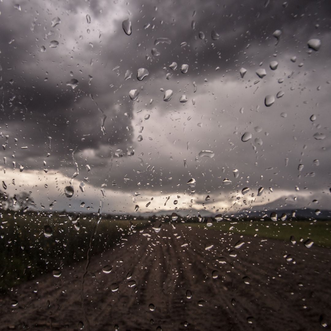 Raindrops cover a window, creating water streaks and droplets across the glass. Beyond the window, an outdoor landscape with fields and distant hills is visible under dark, heavy storm clouds.
